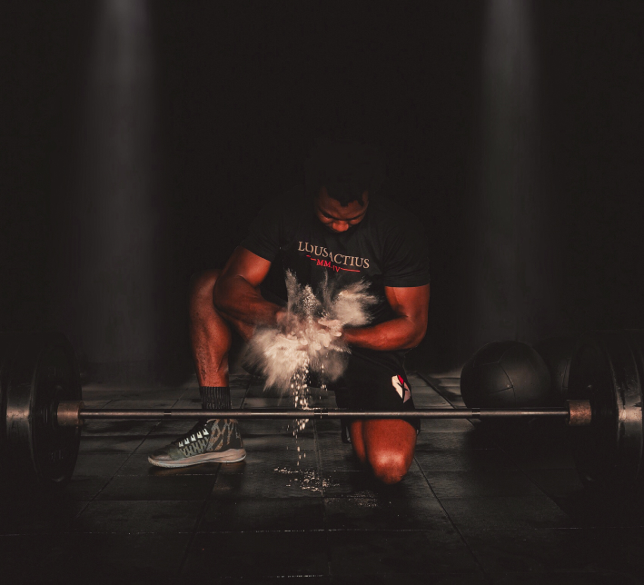 A member kneeling behind a loaded barbell, clapping chalk from his hands, in a Ludus Actius shirt