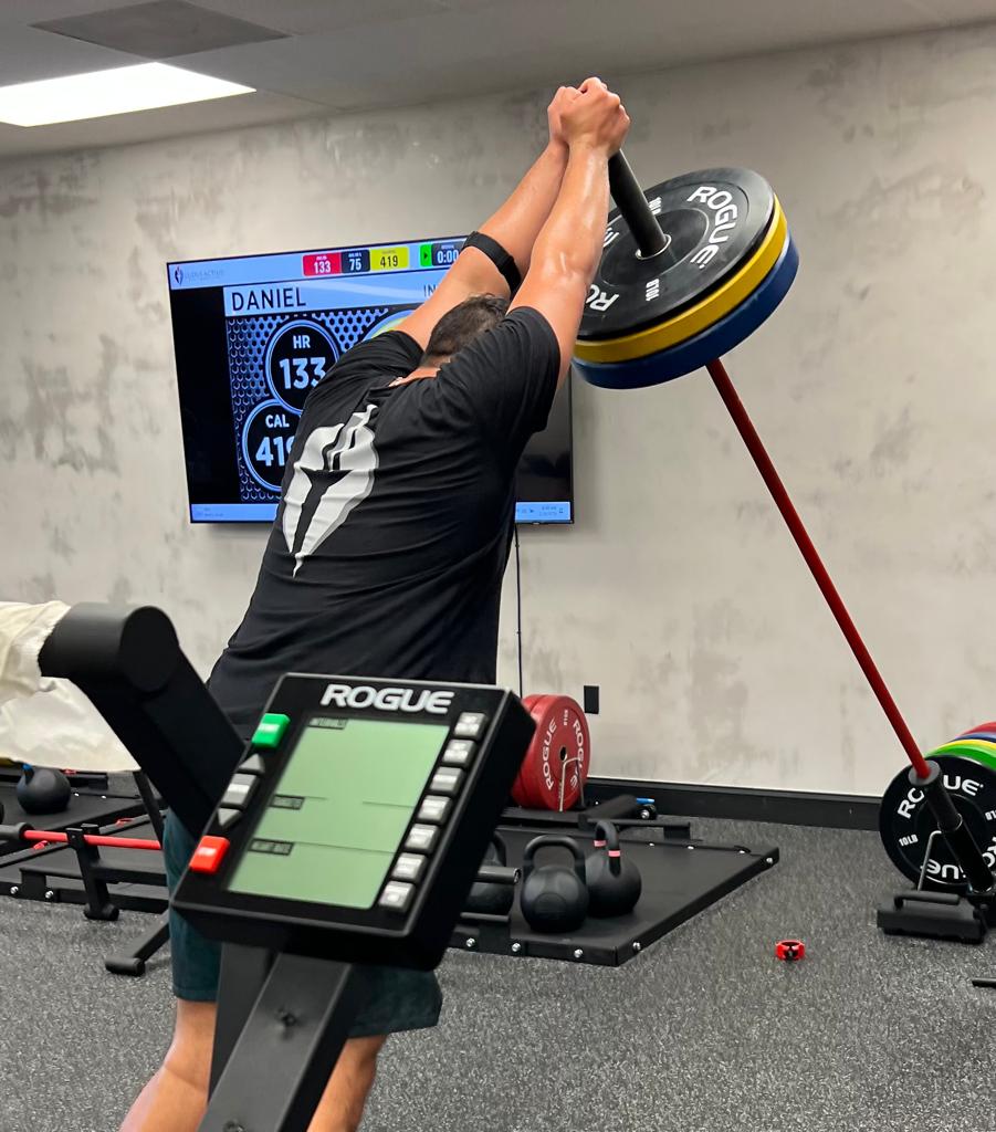 A member pressing a loaded bar overhead, with the heart rate board on the wall showing his numbers