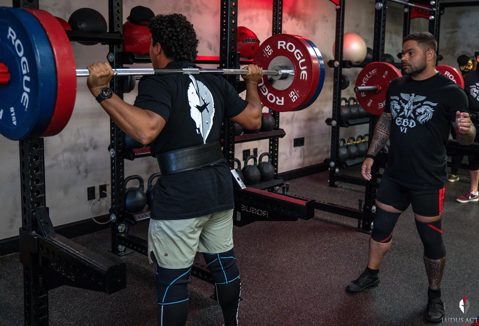 A trainer coaching a member through a back squat in the rack at Ludus Actius
