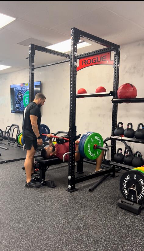 A coach spotting a member on the bench press, with the heart rate board on the wall behind